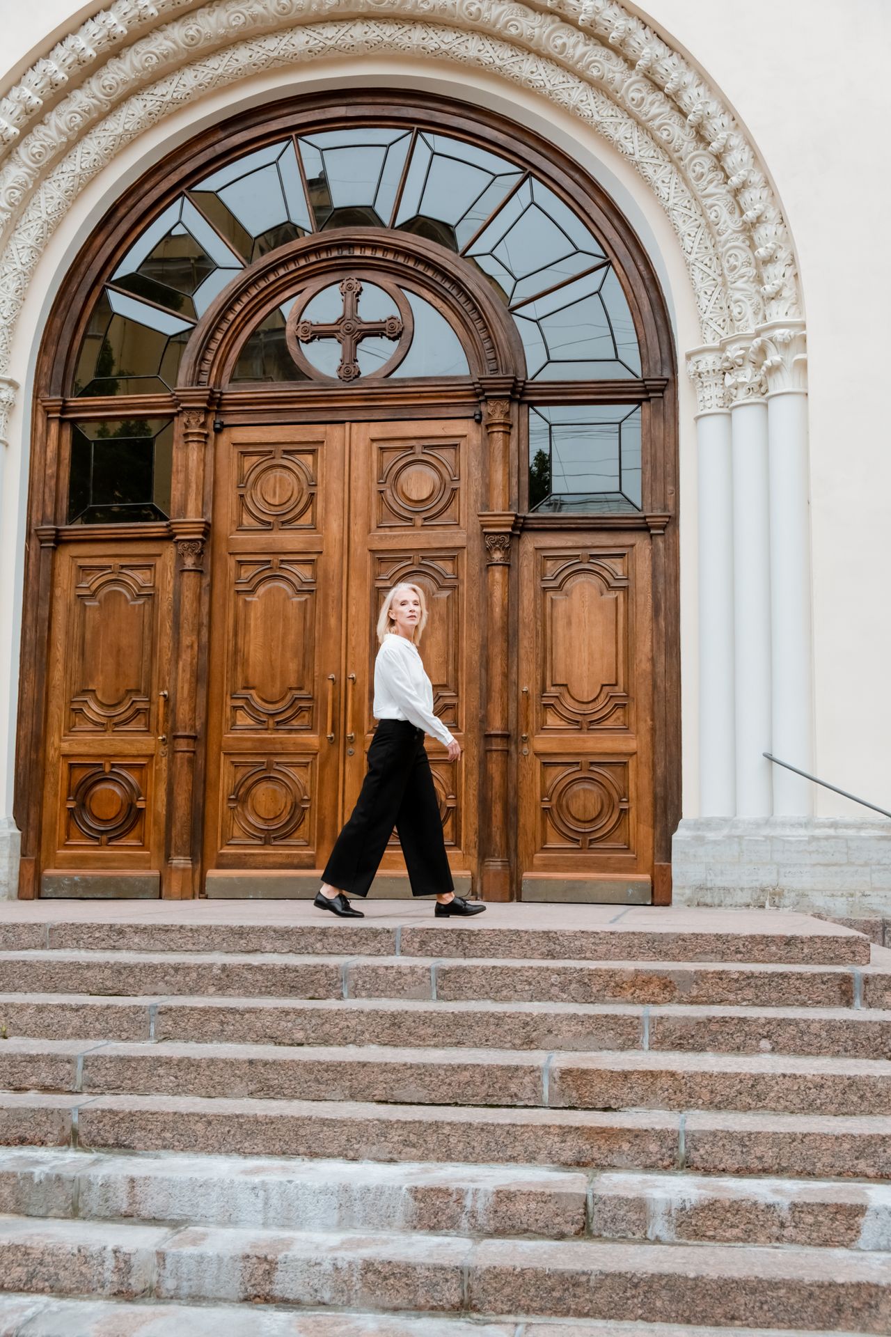 Elegantna žena u crnim hlačama i bijeloj košulji prolazi ispred masivnih povijesnih vrata za moderan poslovni stil