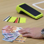 Bankovne kartice i POS uređaj na drvenom stolu uz ruku koja drži euro novčanice