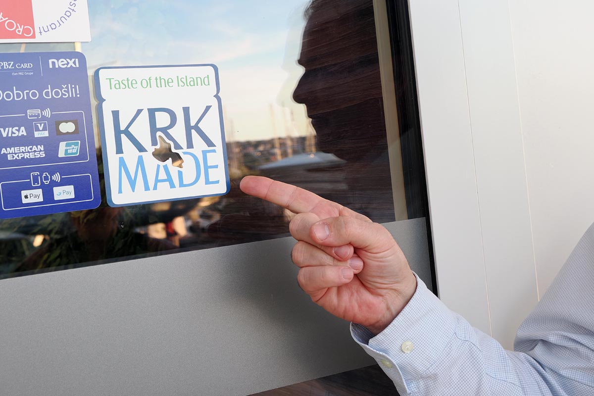 Petar Škarpa pokazuje na plavu Krk Made naljepnicu na staklenim vratima restorana u Puntu