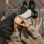 Osoba u bež baloneru, crnim cipelama i trapericama drži cappuccino u krilu