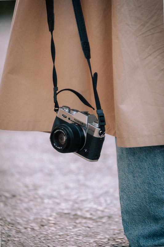 Srebrni Fujifilm fotoaparat na crnom remenu visi preko ramena osobe u dugom bež mantilu i trapericama