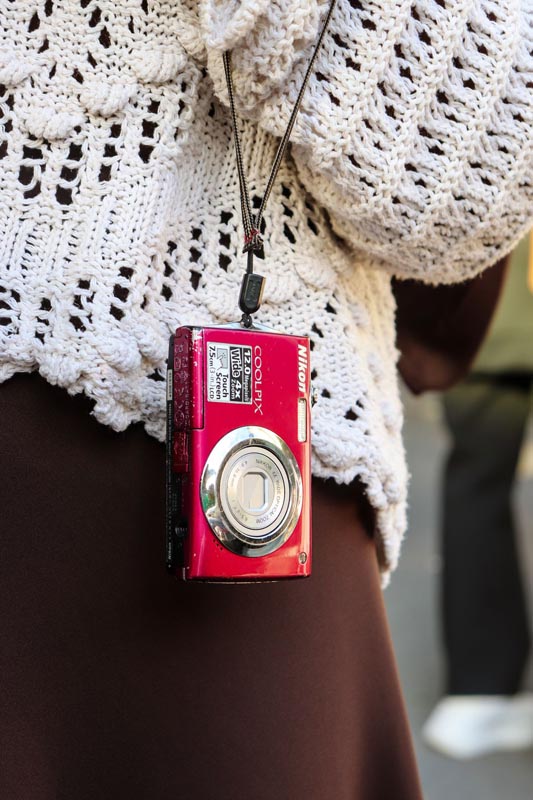 Mali crveni Nikon Coolpix fotoaparat visi na tankoj uzici oko vrata osobe u bijelom pletenom džemperu