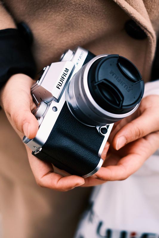 Osoba u bež kaputu drži srebrno-crni Fujifilm X-T200 fotoaparat s poklopcem na objektivu