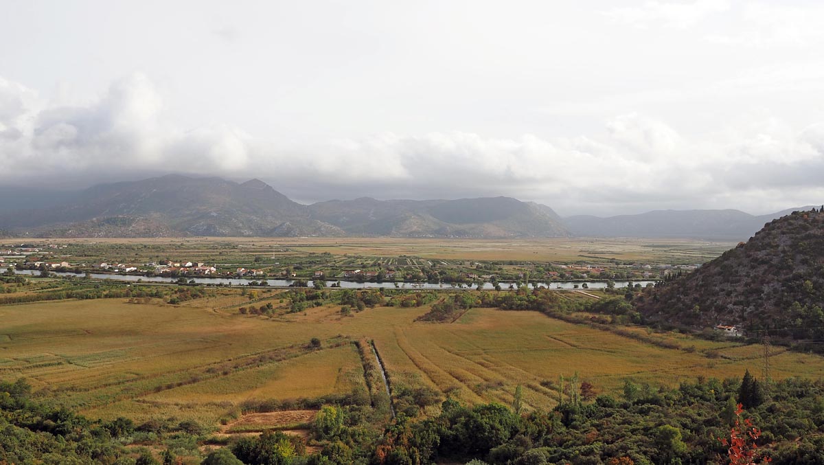 Panoramski pogled na prostranu dolinu Neretve s poljima, rijekom i planinama u pozadini pod oblacima