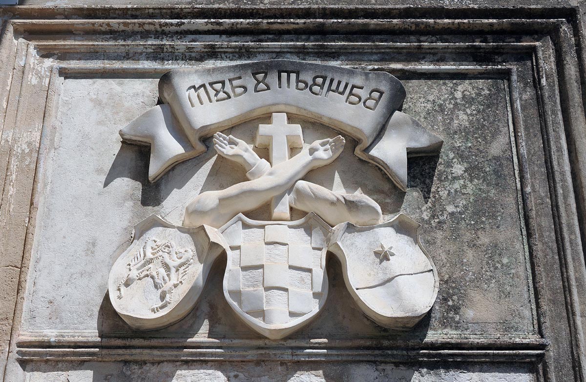 Isklesani franjevački grb s prekriženim rukama, natpisom na glagoljici i povijesnim šahovnicama na zidu