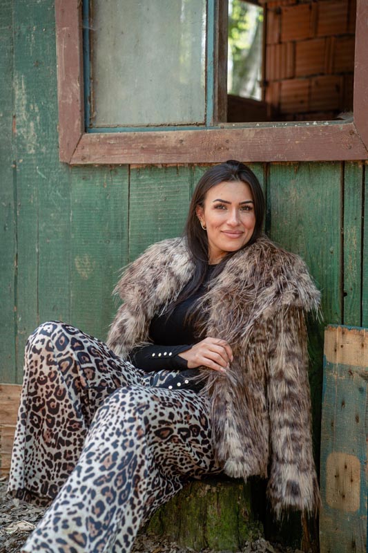 Brineta u pufastoj krznenoj jakni i širokim leopard hlačama sjedi ispred stare zelene drvene kućice