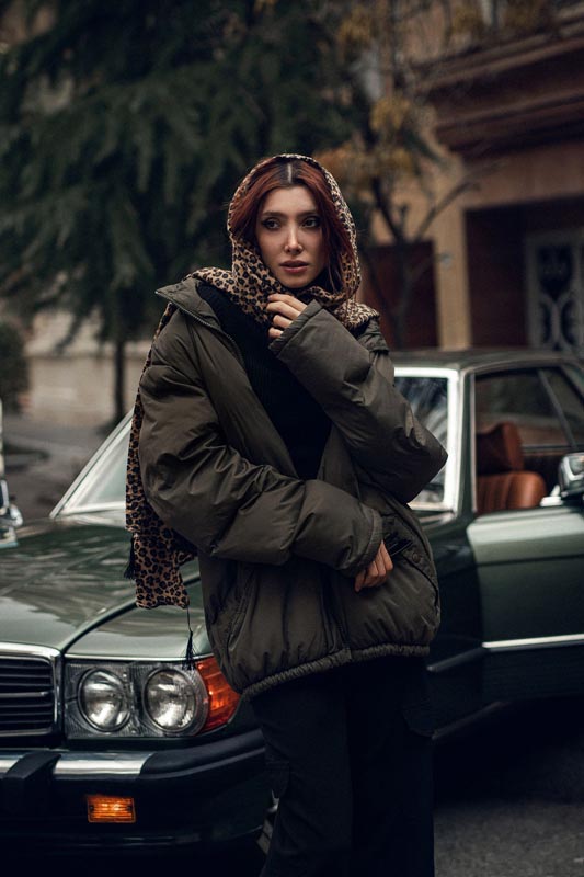 Žena u maslinastozelenoj puffer jakni i s leopard maramom na glavi stoji pokraj zelenog retro automobila