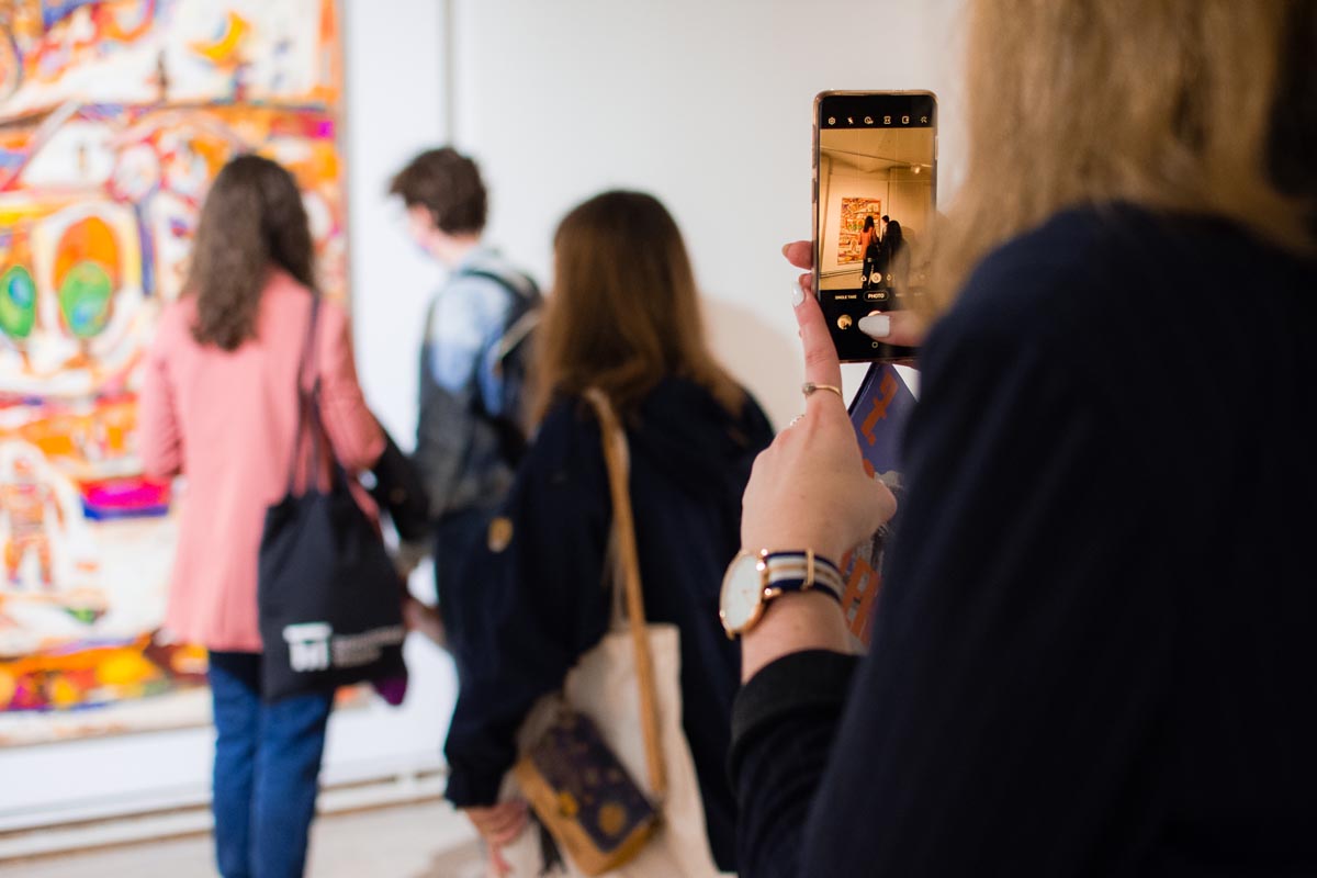 Posjetiteljica mobitelom fotografira izložbu u galeriji dok drugi ljudi razgledavaju šarene slike