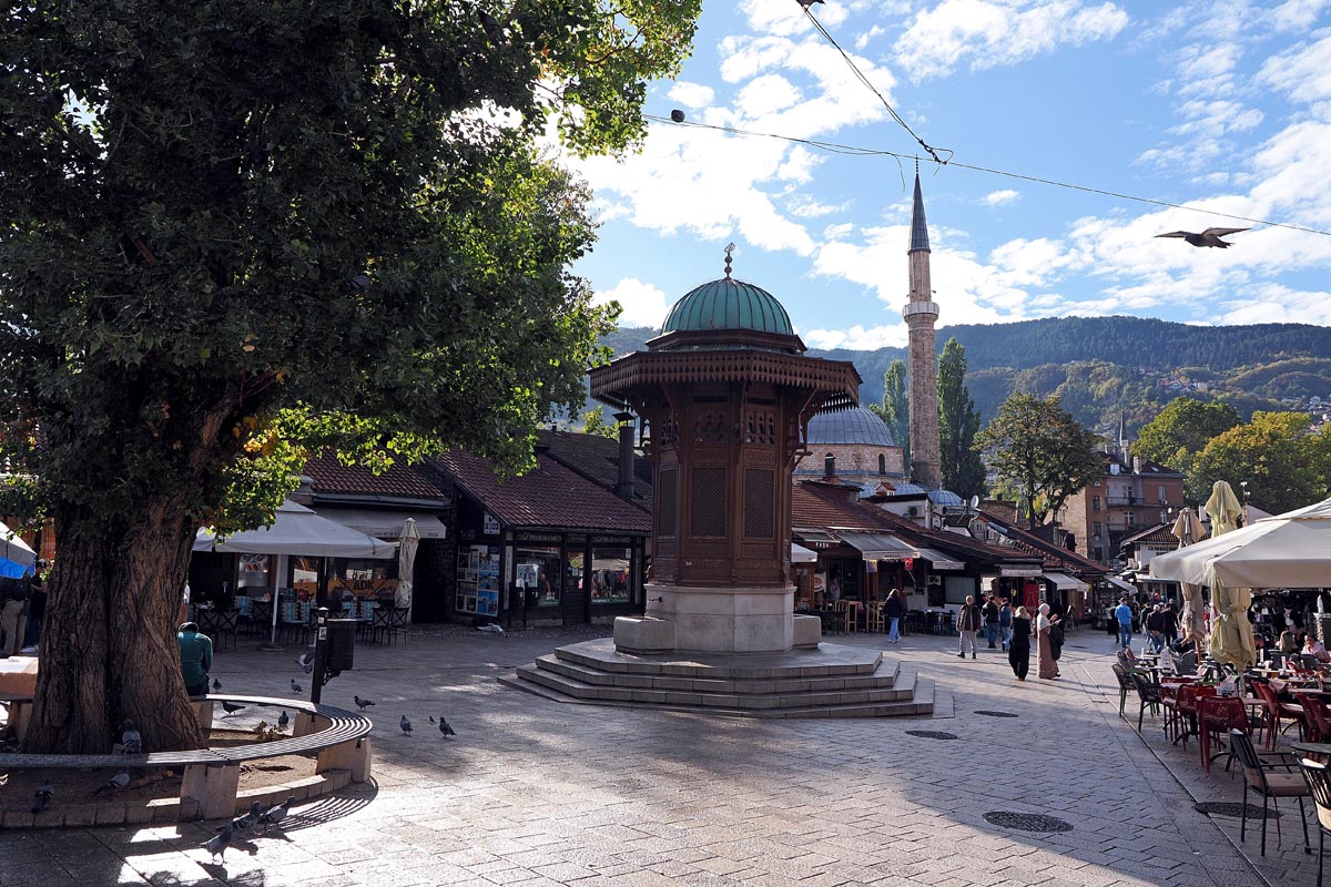 Drveni Sebilj na Baščaršiji u Sarajevu okružen golubovima pod plavim nebom s oblacima