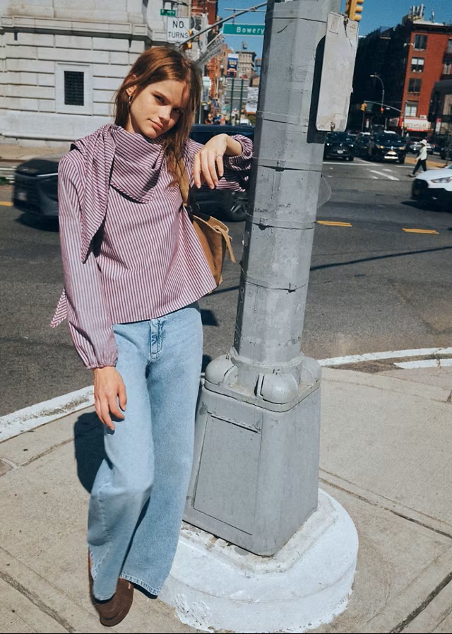 Djevojka u prugastoj Mango košulji i širokim trapericama naslonjena na ulični stup u New Yorku