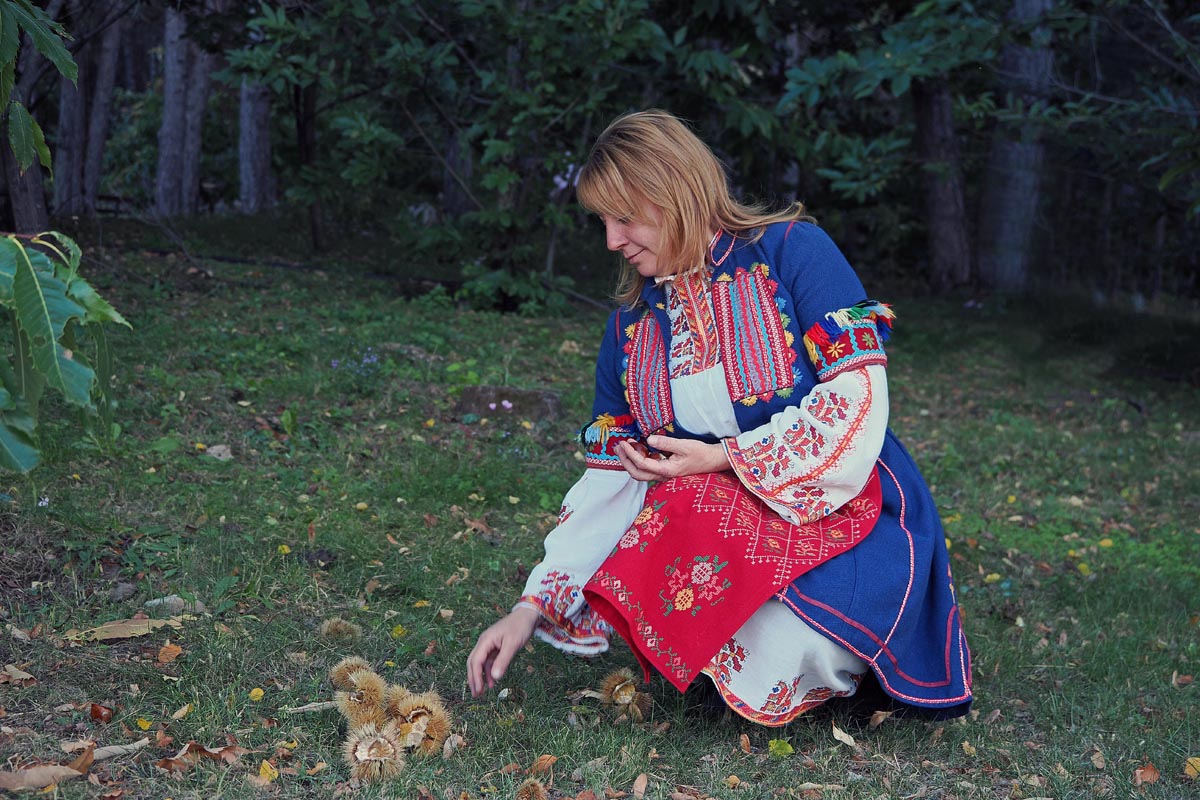 Žena u tradicionalnoj bugarskoj narodnoj nošnji skuplja kestene na travi ispred šume