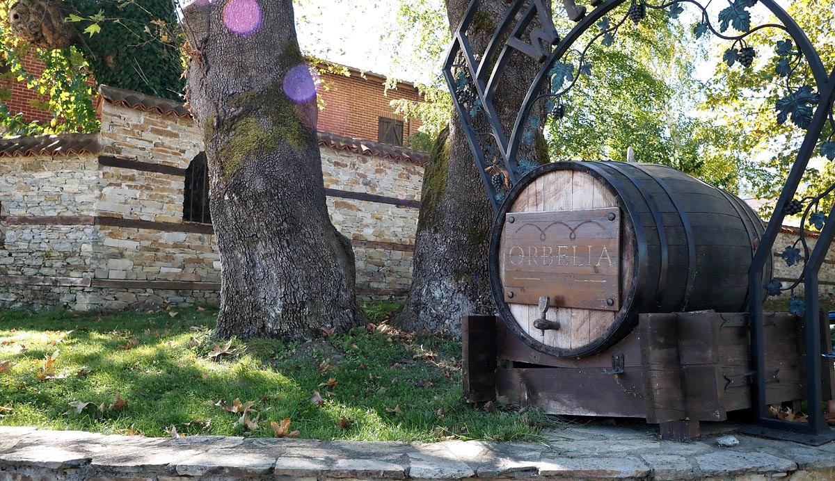 Velika drvena bačva s natpisom Orbelia na postolju u dvorištu vinarije ispred starih kamenih zidova i stabala